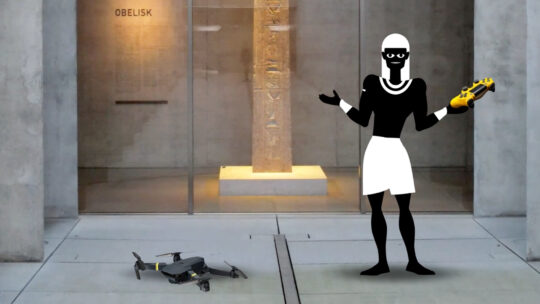 Tjeti steht im Atrium des Ägyptischen Museums und hat fragend die Hände erhoben. In einer Hand hält er eine Fernsteuerung, neben ihm auf dem Boden liegt eine abgestürzte Drohne.