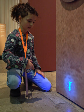 Ein Kind leuchtet mit einer UV-aschenlampe auf eine Wand. Dort ist eine im Licht strahlende Zeichnung einer Maus zu erkennen.