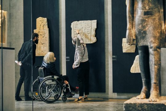 Eine alte Dame in einem Rollstuhl, begleitet von zwei anderen Personen. Alle drei betrachten ein Relief im Museum.
