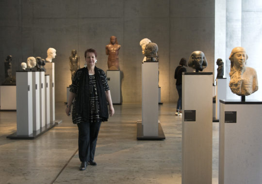 Dr. Sylvia Schoske steht inmitten von altägyptischen Skulpturen im Museum