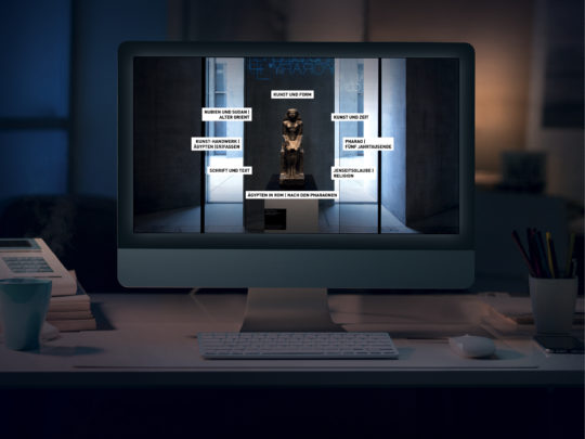 Ein Computerbildschirm steht in einem dunklen Raum, auf dem Bildschirm ist die Startseite der Grand Tour, der digitalen Ausstellung, angezeigt