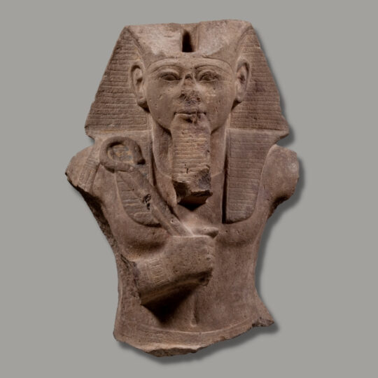 Statuenoberteil von Ramses II. aus hellem Sandstein. Der König trägt ein gestreiftes Kopftuch, in einer Hand hält er vor der Brust Krummstab und Wedel. Beide Arme sind abgebrochen.