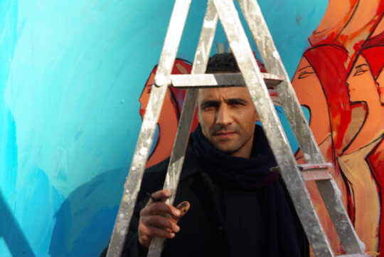 Der Künstler Alaa Awad steht unter einer Leiter vor einem seiner Wandgemälde.