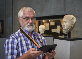 Ein älterer Mann steht vor einem Statuenkopf im Museum und schaut auf den Bidlschrim des MedienGuide
