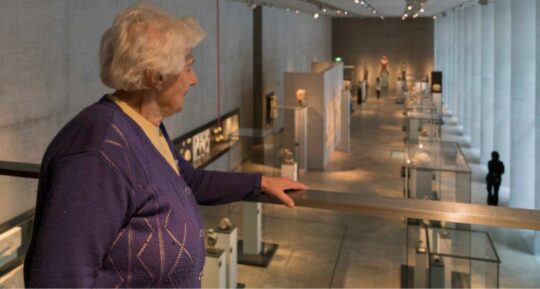 Eine alte Dame steht auf der Galerie des Museums und blickt in den Ausstellungsraum