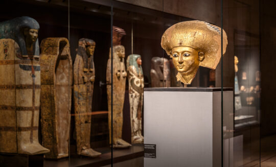 Ausstellungsraum "Jenseitsglaube" - die Goldmaske der Satdjehuti in ihrer Vitrine, auf der linken Seite eine Reihe von Särgen.