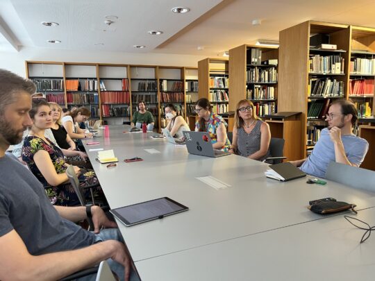 Eine Gruppe von Personen sitzt um einen großen Besprechungstisch in einer Bibliothek.