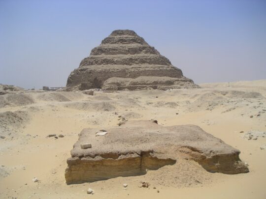 Blick über einen im Sand liegenden Altar auf die Stufenpyramide des Djoser