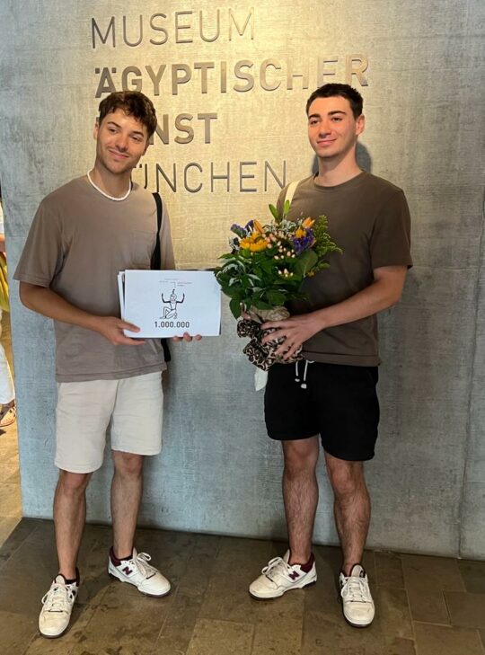 Zwei junge Männer stehen vor einer Betonwand mit dem Namen des Museums. Einer hält einen Blumenstrauß in der Hand, der andere ein Schild mit 1.000.000