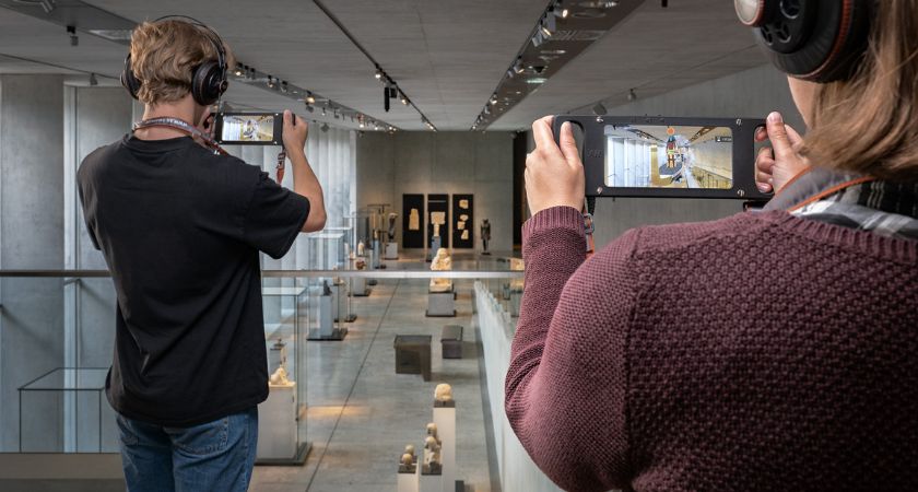 Zwei menschen in Rückanischt stehen auf der Galerie des Museums und halten Mobilgeräte in den Händen. Beide tragen Kopfhörer