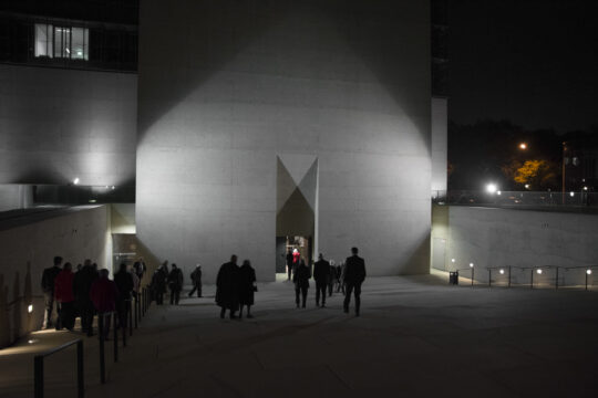 Menschen gehen über die Außentreppe in das Museum. Es ist Nacht, die Portalwand ist beleuchtet.