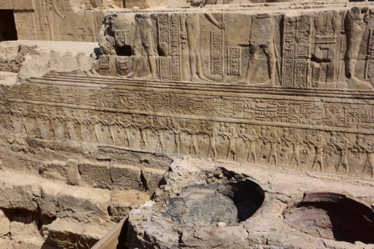 Sockelleiste eines Tempel aus braunem Sandstein. Ganz unten eine Reihe von Gottheiten und darüber eine Reihe Hieroglyphen.