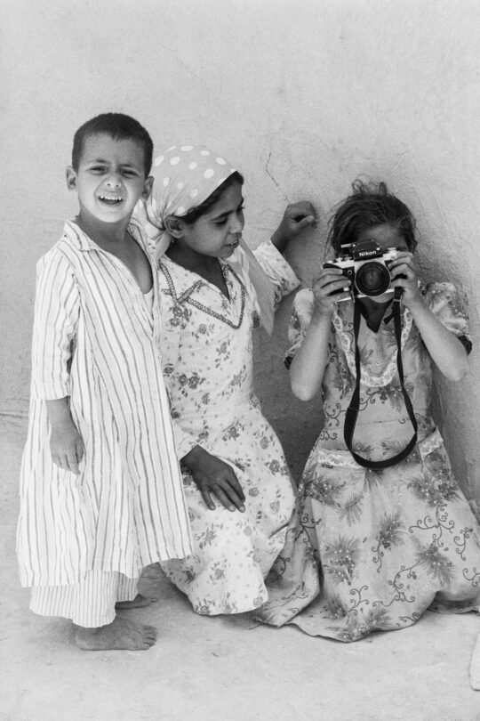 Drei kleine Kinder in traditionell ägyptischer Kleidung. Das linke schaut halb lachend, halb fragend, die betrachtende Person an, das rechte Kind hält eine Kamera.