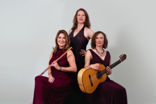 Die drei Mitglieder von Tre Fiamme - drei mittelalte Frauen mit langen braunen Haaren und langen dunkelroten Kleidern. Eine hält eine Querflöte in der Hand, eine andere eine Gitarre.