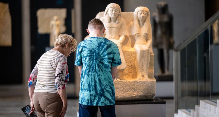Eine ältere Dame und ein Jugendlicher stehen gemeinsam vor einer altägyptischen Statue und betrachten diese.