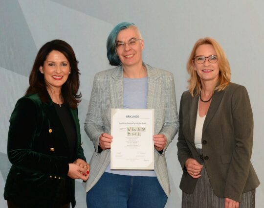 Staatsministerin Michaela Kaniber, Roxane Bicker, Barbara radomski, Geschäftsführerin der Bayern Toursimus Marketing GmbH. Die drei Personen stehen nebeneinander, Roxane Bicker hält die zertifizierungsurkunde in der Hand.