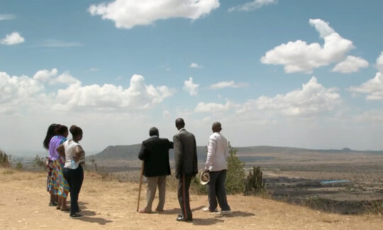 Auf einem flachen Hügel in einer Steppenlandschaft stehen mehrere Personen und schauen in die Ferne.