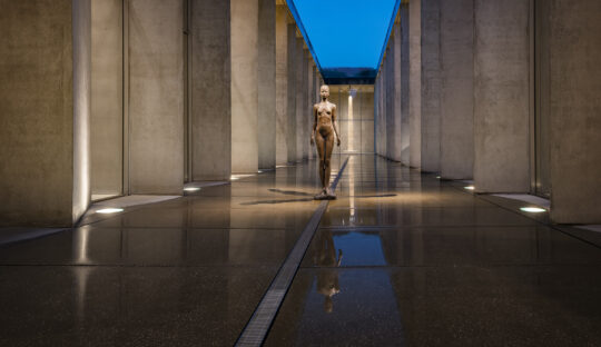 Abendliche Aufnahme im Museum zur blauen Stunde. Blick in das nassgeregnete Atrium, darüber ein tiefblauer Himmel, rechts und links die beleuchteten Betonpfeiler. In der Mitte des Atriums steht eine Bronzeskulptur einer Frau.