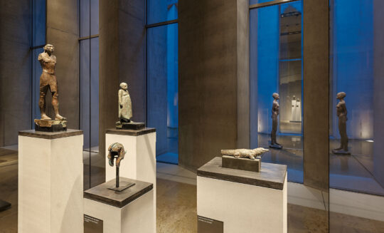 Abendliche Aufnahme im Museum zur blauen Stunde. Im Vordergrund stehen vier Bronzestatuen in einer Vitrine, im Hintergrund sieht man durch die Fenster zwischen den Betonpfeiler blaues Licht.