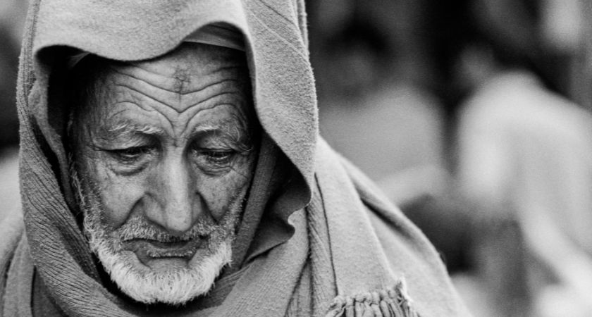 Schwarzweißbild eines alten Mannes mit einem Schal über den Kopf. Er hat ein zerfurchtes Gesicht und einen weißen Bart und schaut halb nach unten