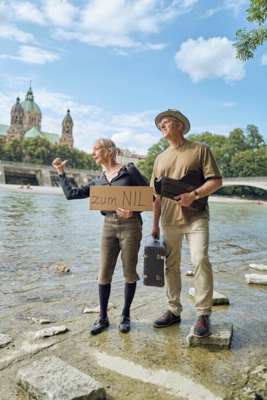 Georg Glasl und Ruth Geiersberger stehen an der Isar. Glasl hält in einer Hand eine Zither, in der anderen einen alten Reisekoffer. Geiersberger hebt den Daumen und hält ein Pappschild, auf dem steht "zum NIL".