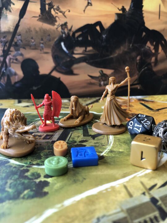 Auf einem bunten Spielbrett stehen ägyptisierende Spielfiguren, eine Ammit, eine Sphinx, ein falkenköpfiger Krieger und ein Dämonenbeschwörer, daneben liegen drei Würfel und einige bunte Spielechips.