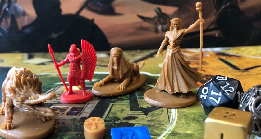 Auf einem bunten Spielbrett stehen ägyptisierende Spielfiguren, eine Ammit, eine Sphinx, ein falkenköpfiger Krieger und ein Dämonenbeschwörer, daneben liegen drei Würfel und einige bunte Spielechips.