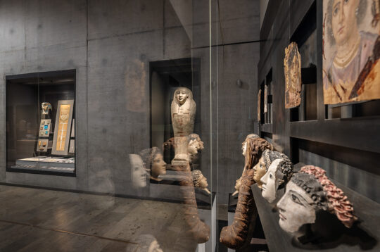 Blick auf Stuckmasken und einen Steinsarkophag in einer Vitrine