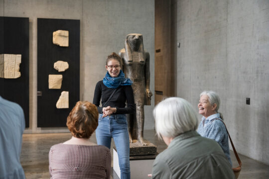 Eine Gruppe älterer Menschen lauscht einer jungen Kulturvermittlerin im Museum