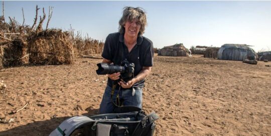 Michael Martin kniet im Sand einer Steppe und hält eine Kamera in der Hand, vor ihm eine Fototasche.