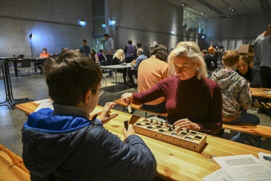 Zwei Personen sitzen an einem Holztisch, zwischen ihnen eine Brettspielbox aus Holz mit altägyptischen Hieroglyphen.