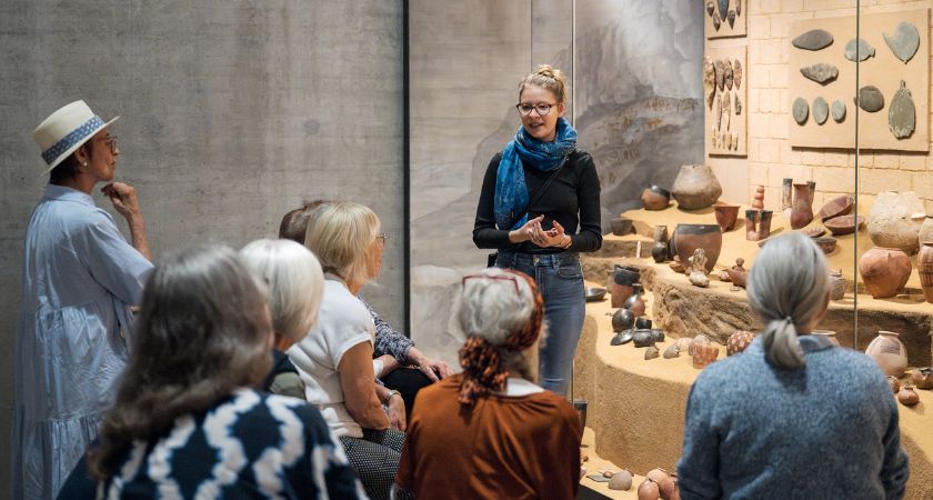 Eine Gruppe älterer Damen lauscht einer jungen Führungskraft. Alle haben sich vor einer Vitrine versammelt.