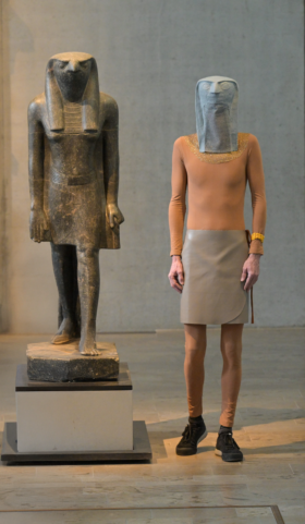 Ein als Horus verkleideter Mensch steht neben der Statue des Gottes Horus im Museum