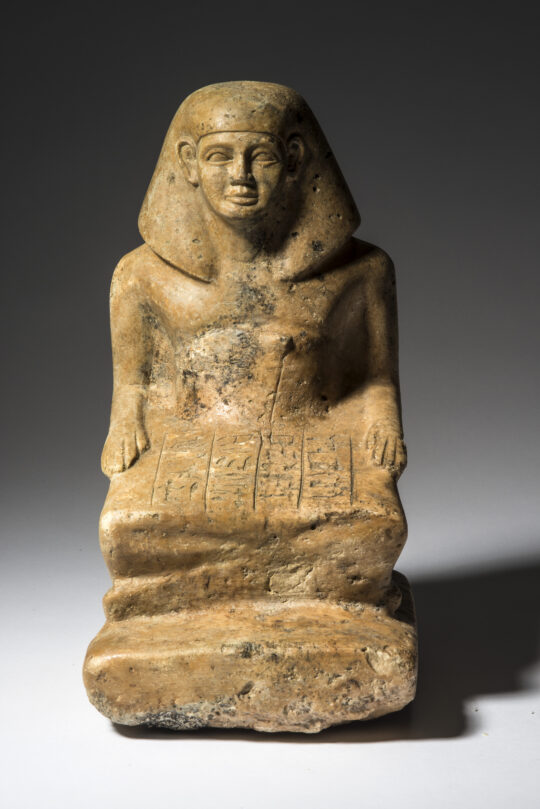 Altägyptische Statue eines Lesenden aus hellgelbem Stein. Ein Mann sitzt mit untergeschlagenen Beinen und hält darauf einen ausgerollten Papyrus