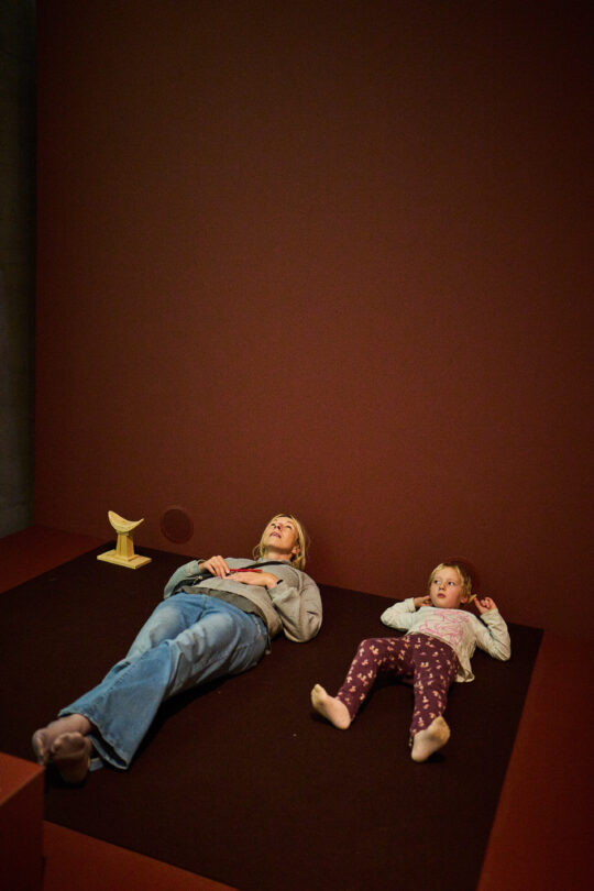 Eine Frau und ein Kind liegen auf einem Teppichboden, den Kopf in eine Kopfstütze gelegt.