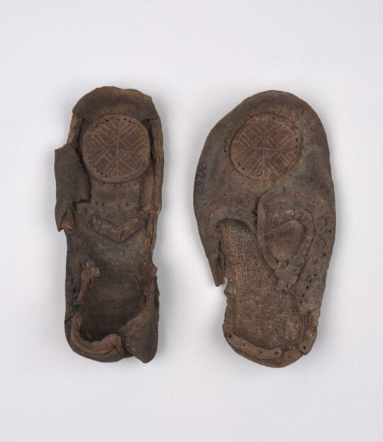 Ein paar kleine lederne Kinderschuhe in geschlossener Form mit einer Art Rosette auf dem vorderen Schuhteil