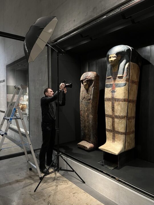 Ein Mann steht vor einer offenen Vitrine im Museum und fotografiert Särge.