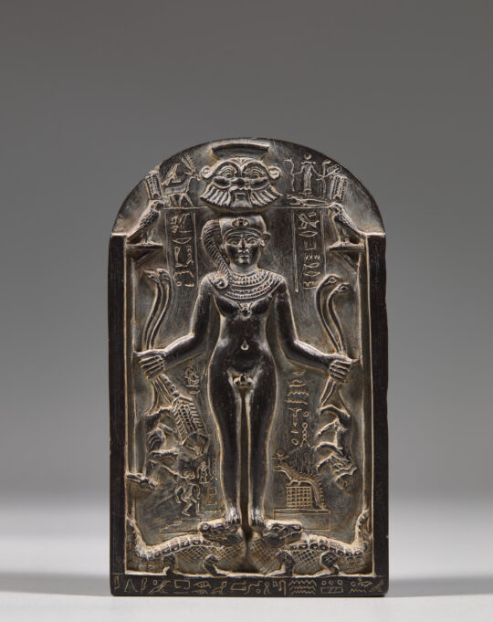 Eine kleine Stele, oben halb gerundet, aus schwarzem Stein. In der Mitte in erhabenem Relief eine Abbildung des Gottes Horus in Kindgestalt, der Schlangen, Skorpione und Löwen in den Händen hält und auf zwei Krokodilen steht.