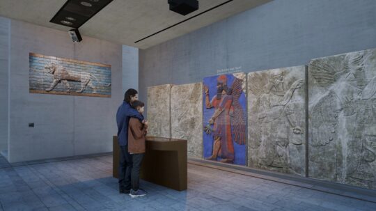 Zwei menschen stehen vor einer Medienstation die ein assyrisches Relief an der gegenüberliegenden Wand virtuell einfärbt