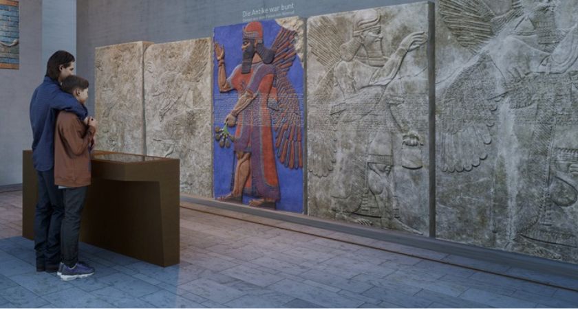 Computergenerierte Ansicht des Raumes Alter Orient. Vor der Medienstation stehen zwei Menschen, eines der Reliefs an der Wand ist bunt beleuchtet.