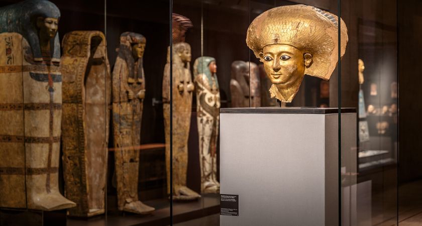 Blick in den Raum Jenseitsglaube, zentral im Bildfeld die goldene Sargmaske der Satdjehuti, im Hintergrund in einer Vitrine menschengestaltige Särge