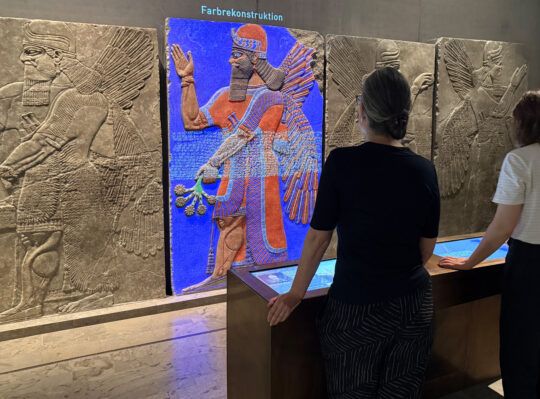 Zwei Personen stehen an einer Medinstation. Im Hintergrund ist eines der assyrischen Reliefs bunt beleuchtet.