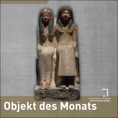 Frontalaufsicht auf eine altägyptische Gruppenstatue, rechts sitzt ein Mann mit langem Gewand und einer Perücke, deren Haare über seine Schultern fallen, neben ihm eine Frau in einem langen, enganliegenden Kleid, deren Perückenhaare bis auf ihre Brüste fallen. Die Statue ist aus hellem Stein, Reste von Bemalung haben sich erhalten, die Haare sind schwarz, die Gesichter rötlich-braun.