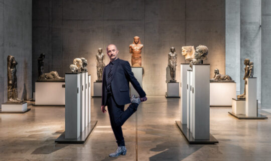 Ein Mann steht zwischen den Statuen in der Ausstellung, er hält seinen linken Fuß mit der linken Hand gegriffen und zieht ihn hoch.