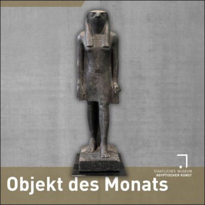 Statue eines falkenköpfigen, menschengestaltigen Gottes aus schwarzem Stein vor betonhintergrund. Unten steht in weißer Schrift auf olivfarbenem Grund: Objekt des Monats